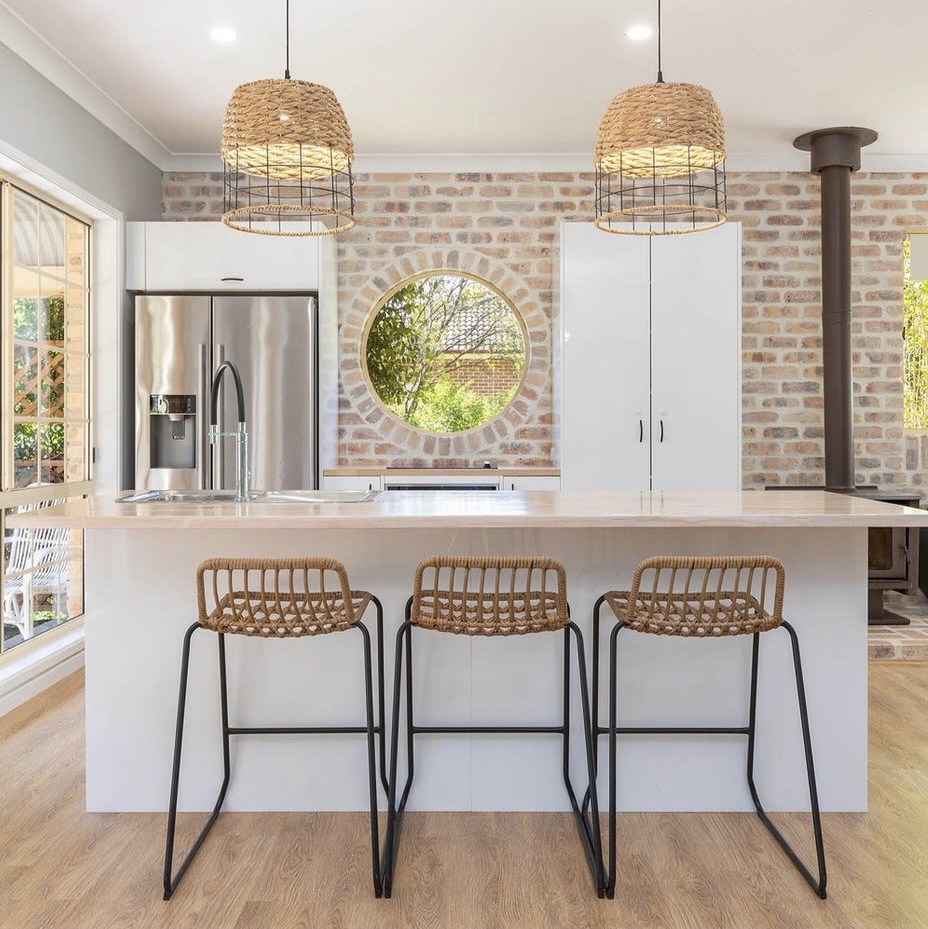 Country kitchen with recycled brick feature wall, round window and two woven rattan pendant lights over a stone island