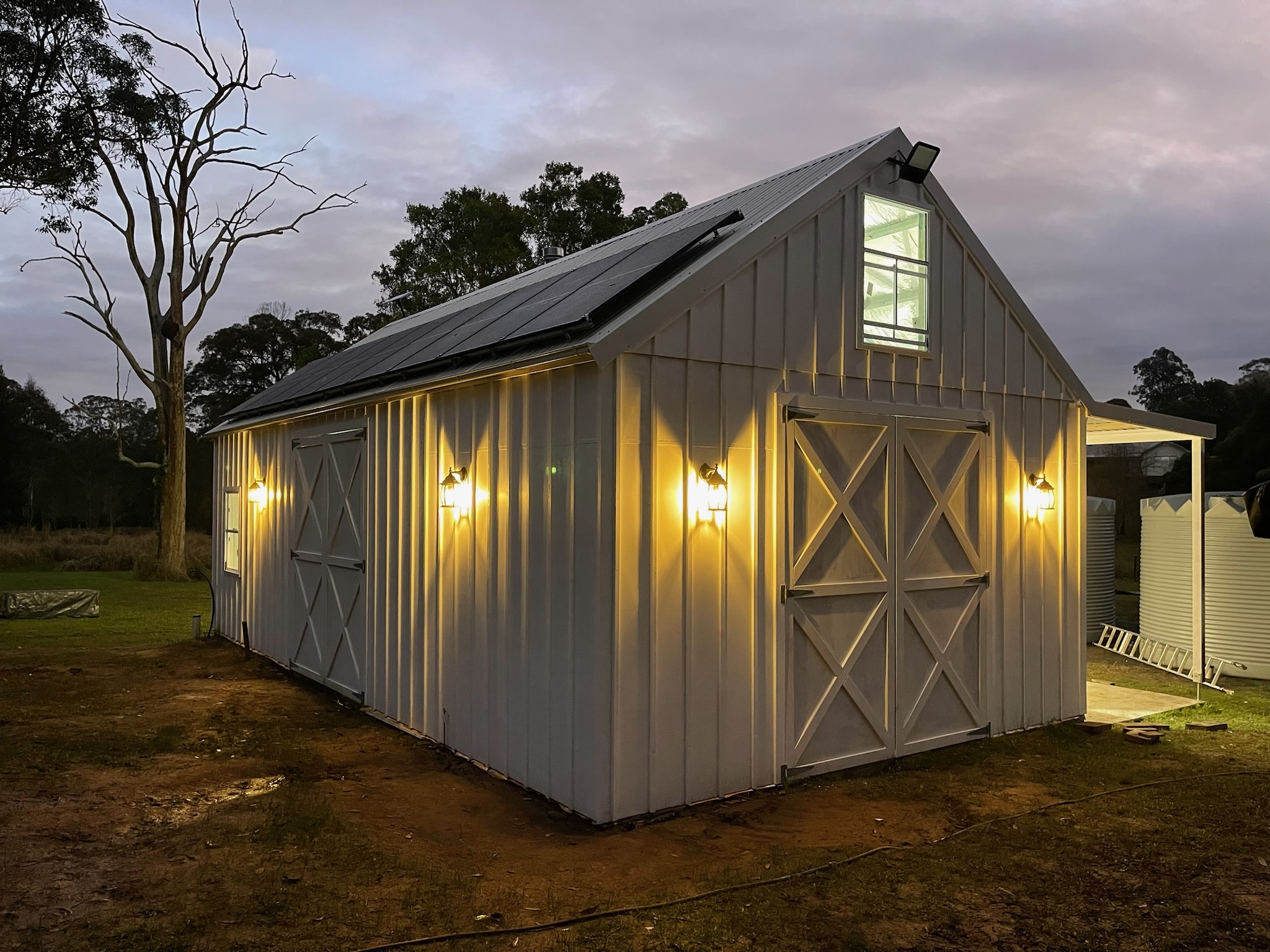 Barn style shed at dusk with five wall lanterns lit along the side and solar panels on the roof