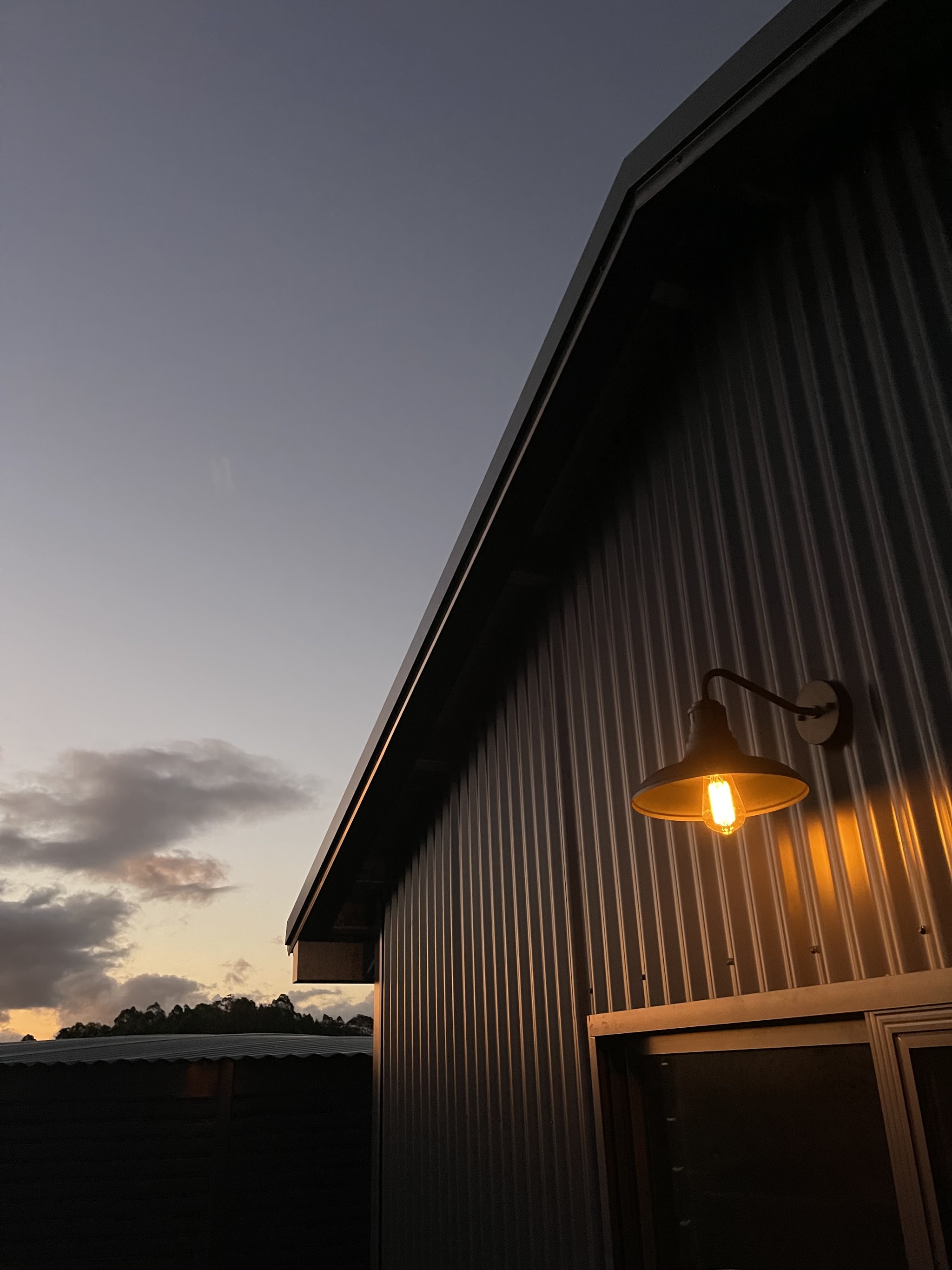 Dark corrugated iron barn at dusk with a single warm gooseneck light glowing against a violet sky